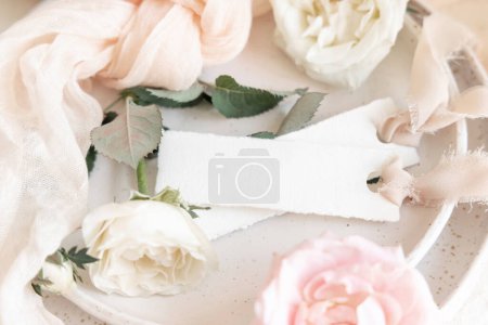 Foto de Tarjeta en blanco horizontal cerca de tela de tul rosa claro y flores de crema en el plato de cerca, espacio de copia. Papelería de boda maqueta. Tarjetas románticas y decoración pastel - Imagen libre de derechos