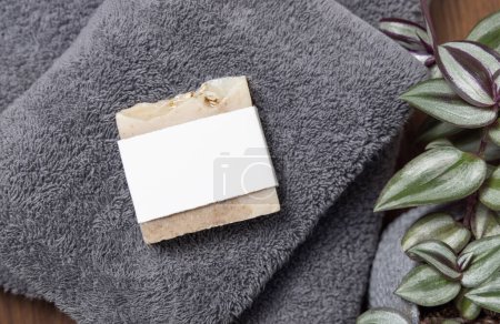 Seifenbar mit leerem Etikett auf grau gefalteten Handtüchern in der Nähe grüner Pflanze auf brauner Holzarbeitsplatte im Badezimmer, Draufsicht, Markenverpackungsattrappe. Natürliches handgemachtes Hygieneprodukt