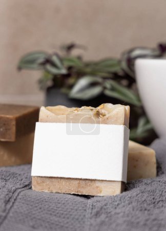 Barra de jabón beige con etiqueta en blanco cerca de toallas dobladas grises, planta verde y recipiente en el baño, primer plano, maqueta de embalaje de marca. Producto natural de higiene artesanal