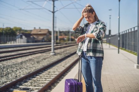 Téléchargez les photos : Femme inquiète regardant son horloge debout avec une valise sur une gare. - en image libre de droit