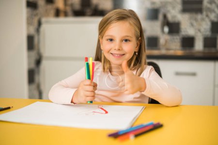 Foto de Hermosa niña mostrando el pulgar hacia arriba mientras disfruta dibujando en su casa. - Imagen libre de derechos