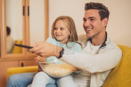 Téléchargez les photos : Père et fille aiment regarder la télévision et manger des cors pop ensemble à la maison. - en image libre de droit