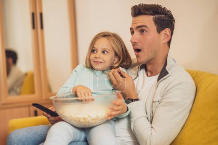 Téléchargez les photos : Surpris père et fille regardant la télévision et mangeant des cors pop ensemble à leur maison. - en image libre de droit