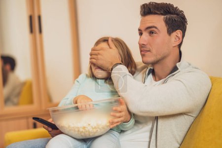 Téléchargez les photos : Père couvrant les yeux de sa fille tout en regardant la télévision et en mangeant des cors pop ensemble à la maison. - en image libre de droit