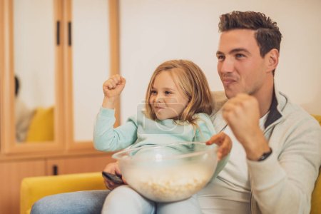 Téléchargez les photos : Père et fille excités aiment regarder la télévision et manger des cors pop ensemble à leur maison. - en image libre de droit