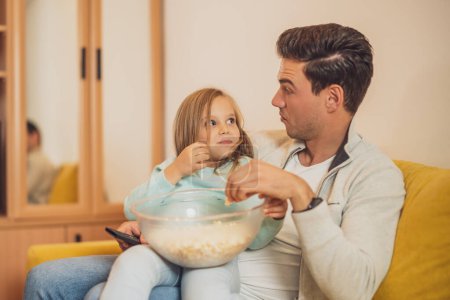 Téléchargez les photos : Surpris père et fille regardant la télévision et mangeant des cors pop ensemble à leur maison. - en image libre de droit