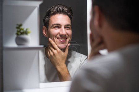 Téléchargez les photos : Portrait of man looking himself in a mirror while standing in bathroom. - en image libre de droit