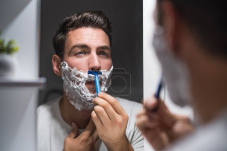 Photo for Man shaving  beard while looking himself in the mirror. - Royalty Free Image