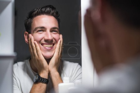 Photo for Man is applying aftershave while looking himself in the mirror. - Royalty Free Image