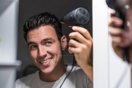 Téléchargez les photos : Man using hair dryer while looking himself in the mirror - en image libre de droit