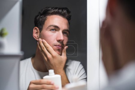 Photo for Man is applying aftershave while looking himself in the mirror. - Royalty Free Image