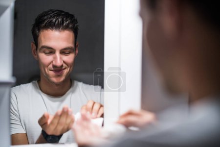 Photo for Man is applying aftershave while standing  in front of mirror. - Royalty Free Image