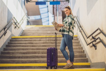 Téléchargez les photos : Femme heureuse avec valise regardant la carte tout en se tenant debout sur les escaliers de la gare. - en image libre de droit