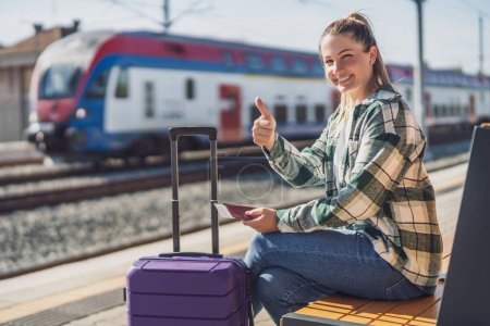 Mujer feliz mostrando el pulgar hacia arriba y sosteniendo el boleto mientras está sentado en el banco en la estación de tren.