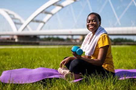 Téléchargez les photos : Heureuse femme sportive tenant des poids tout en étant assise sur un tapis d'exercice extérieur. - en image libre de droit