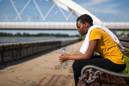 Foto de Mujer agotada después de hacer ejercicio bebiendo agua mientras está sentada en el banco. - Imagen libre de derechos