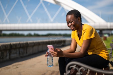Téléchargez les photos : Femme sportive utilisant un téléphone portable tout en se reposant après l'exercice sur le banc et l'eau potable. - en image libre de droit