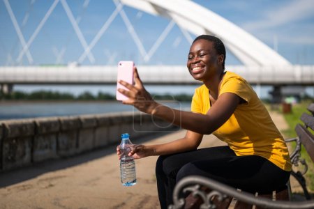 Téléchargez les photos : Femme sportive prenant selfie tout en se reposant après l'exercice sur le banc et l'eau potable. - en image libre de droit