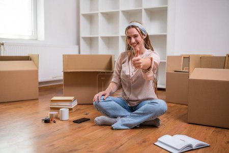 Photo for Modern ginger woman with braids showing thumb up while  moving into new apartment. - Royalty Free Image
