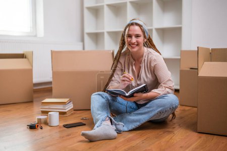 Photo for Modern ginger woman with braids writing in notebook while moving in new home. - Royalty Free Image