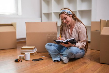 Téléchargez les photos : Femme roux moderne avec des tresses écrivant dans un cahier tout en déménageant dans une nouvelle maison. - en image libre de droit