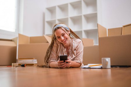 Téléchargez les photos : Femme roux moderne avec des tresses en utilisant le téléphone tout en déménageant dans son nouvel appartement. - en image libre de droit