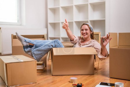 Téléchargez les photos : Femme heureuse montrant pouce vers le haut et la clé de sa nouvelle maison tout en étant assis dans la boîte et en s'amusant. - en image libre de droit