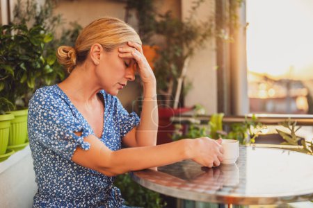 Téléchargez les photos : Triste femme ayant mal à la tête et buvant du café assis sur son balcon. - en image libre de droit