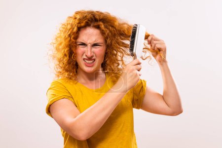 Téléchargez les photos : Femme roux en colère tenant une brosse à cheveux et peignant ses cheveux bouclés. - en image libre de droit