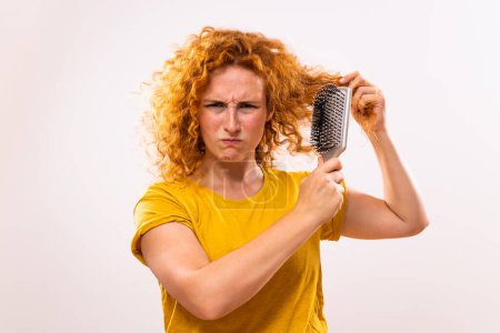 Téléchargez les photos : Femme roux en colère tenant une brosse à cheveux et peignant ses cheveux bouclés. - en image libre de droit