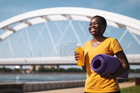 Photo for Sporty woman drinking energy drink and holding  exercise mat. - Royalty Free Image