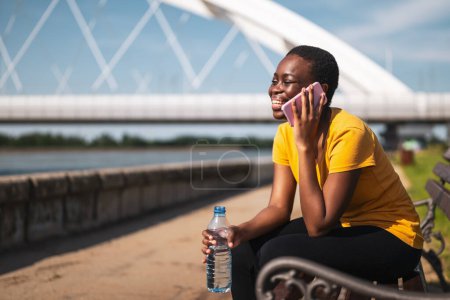 Téléchargez les photos : Femme sportive utilisant un téléphone portable tout en se reposant après l'exercice sur le banc et l'eau potable. - en image libre de droit