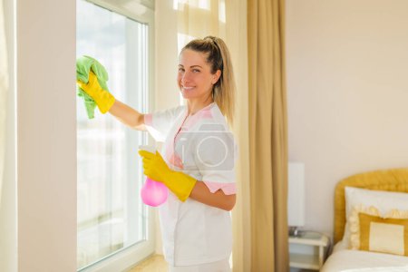 Foto de Imagen de la hermosa criada del hotel limpiando ventanas en una habitación. - Imagen libre de derechos