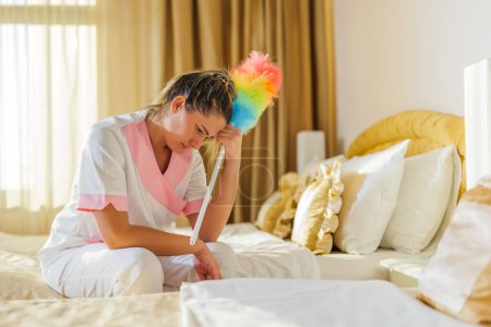 Téléchargez les photos : Image de triste femme de chambre d'hôtel surmenée assise sur le lit dans une chambre. - en image libre de droit
