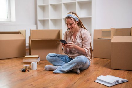 Téléchargez les photos : Femme roux moderne avec des tresses en utilisant le téléphone tout en déménageant dans son nouvel appartement. - en image libre de droit