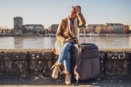 Foto de Turista mujer preocupada con maleta y teléfono perdido en una ciudad sentada junto al río y pensando. Imagen tonificada. - Imagen libre de derechos