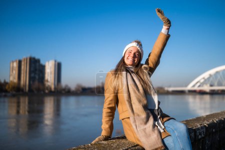 Foto de Hermosa mujer en ropa de abrigo disfruta descansando junto al río en un día soleado de invierno. - Imagen libre de derechos