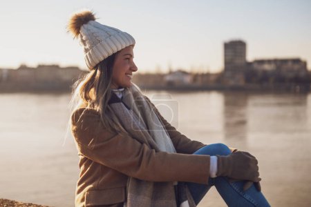 Téléchargez les photos : Belle femme en vêtements chauds aime se reposer au bord de la rivière par une journée d'hiver ensoleillée. Image tonique. - en image libre de droit
