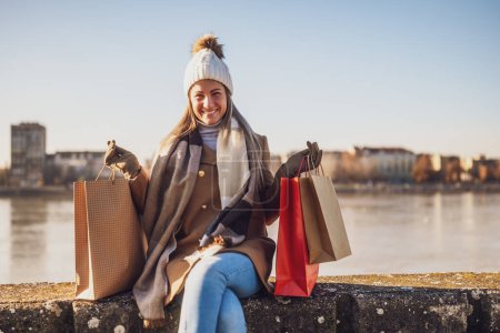 Photo for Happy woman in warm clothing with shopping bags enjoys sitting  by river on sunny winter day. Toned image. - Royalty Free Image