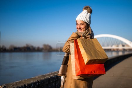 Téléchargez les photos : Femme heureuse en vêtements chauds avec des sacs à provisions jouit debout au bord de la rivière sur une journée d'hiver ensoleillée. - en image libre de droit