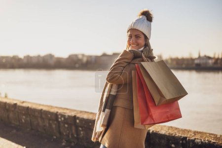 Foto de Mujer feliz en ropa de abrigo con bolsas de compras disfruta de pie junto al río en el soleado día de invierno. Imagen tonificada. - Imagen libre de derechos