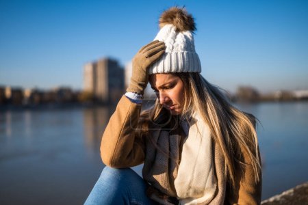 Foto de Mujer preocupada en ropa de abrigo con dolor de cabeza y sentada junto al río en un día soleado de invierno. - Imagen libre de derechos