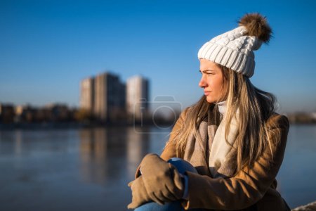 Photo for Sad woman in warm clothing sitting by the river on a sunny winter day. - Royalty Free Image