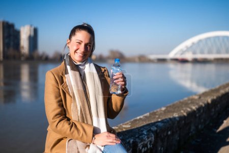 Téléchargez les photos : Femme aime boire de l'eau tout en étant assis au bord de la rivière par une journée d'hiver ensoleillée. - en image libre de droit