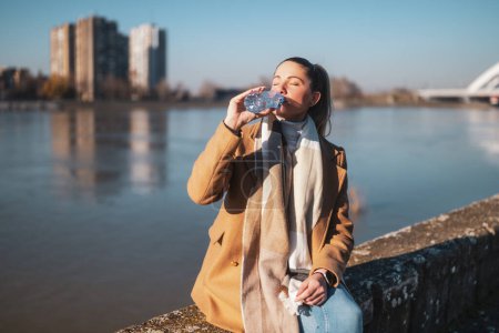 Téléchargez les photos : Femme aime boire de l'eau tout en étant assis au bord de la rivière par une journée d'hiver ensoleillée. Image tonique. - en image libre de droit