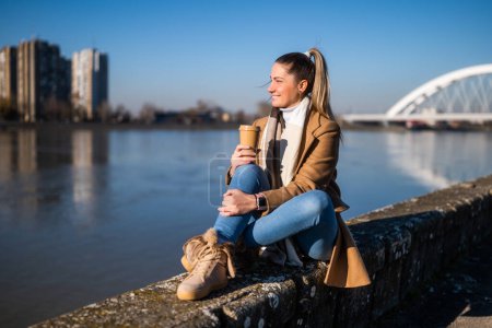 Foto de Hermosa mujer en ropa de abrigo disfruta de beber café y descansar junto al río en un día soleado de invierno. - Imagen libre de derechos