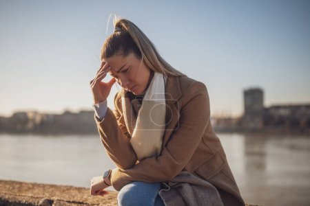 Téléchargez les photos : Femme inquiète ayant mal à la tête tout en étant assis près de la rivière. Image tonique. - en image libre de droit