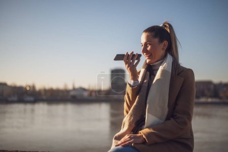 Téléchargez les photos : Belle femme en vêtements chauds parlant sur un téléphone portable et aime se reposer au bord de la rivière par une journée d'hiver ensoleillée. Image tonique. - en image libre de droit