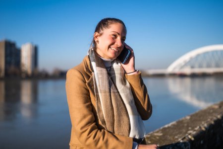 Téléchargez les photos : Belle femme en vêtements chauds parlant sur un téléphone portable et aime se reposer au bord de la rivière par une journée d'hiver ensoleillée. - en image libre de droit