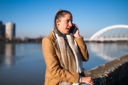 Téléchargez les photos : Femme surprise parlant sur un téléphone portable en vêtements chauds et se reposant au bord de la rivière par une journée d'hiver ensoleillée. - en image libre de droit